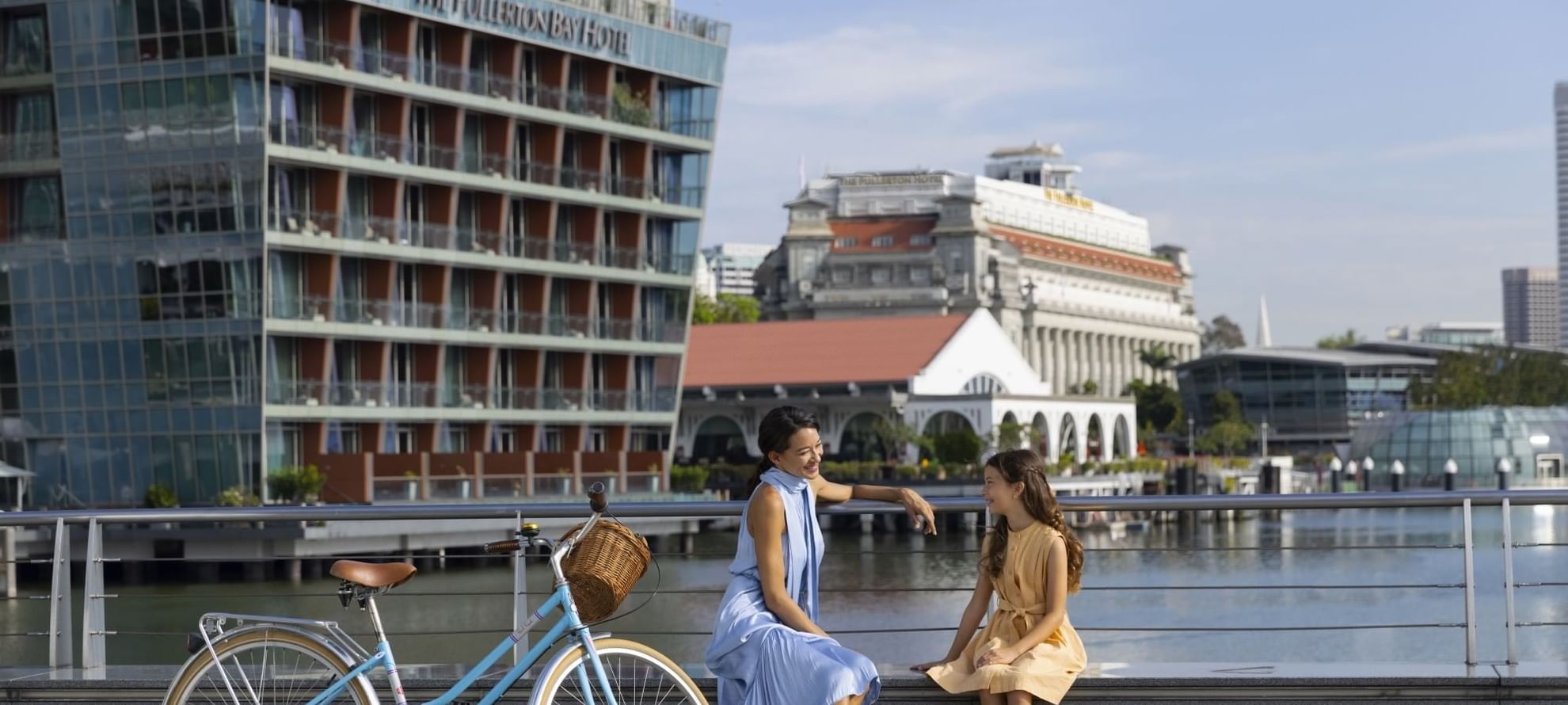 Two people sitting by a riverside, with a bicycle and The Fullerton Hotel Singapore in the background